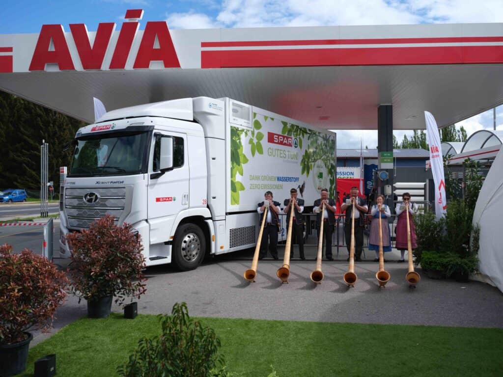 A group of people play alphorns in front of a truck at an AVIA gas station with plants and promotional banners visible.