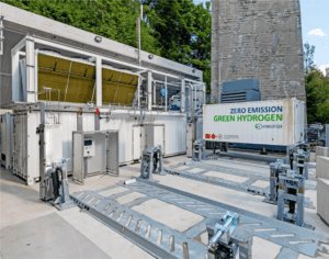 Outdoor green hydrogen production facility with equipment, storage containers, and a sign reading "Zero Emission Green Hydrogen" near a building and trees in the background.