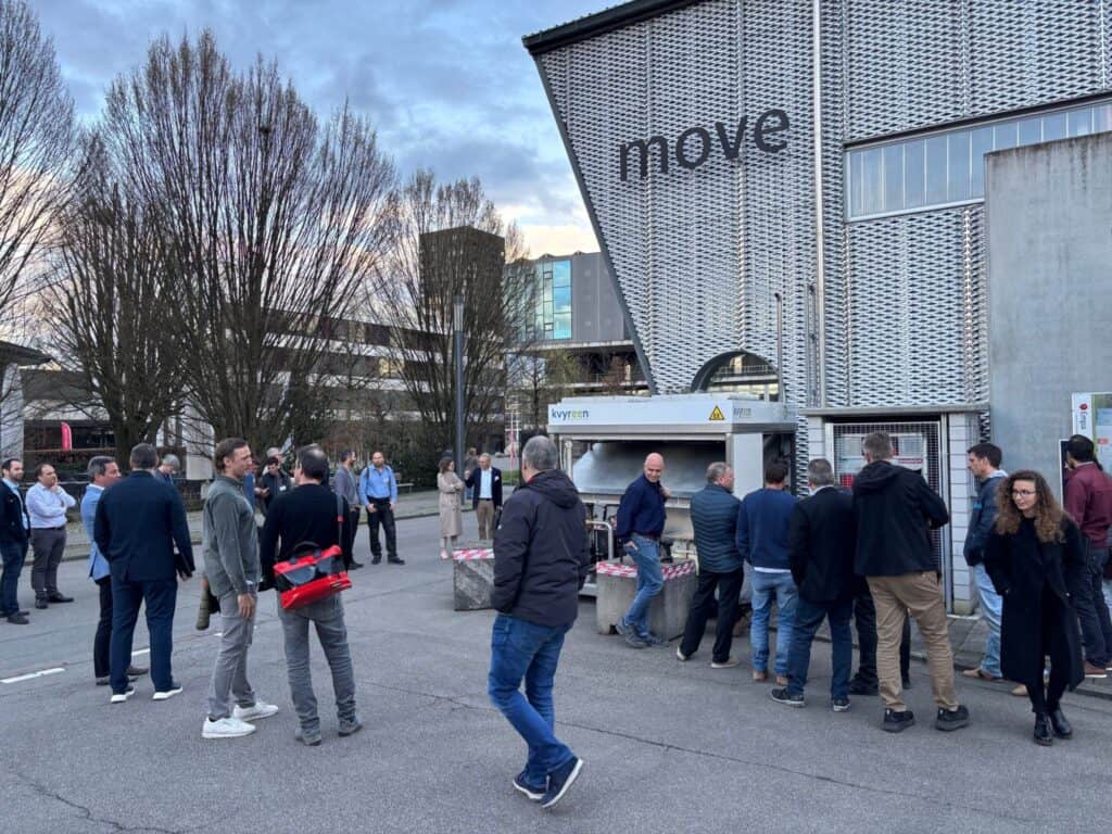 Menschen versammeln sich vor einem modernen Gebäude mit der Aufschrift „Move“, stehen und unterhalten sich in der Nähe eines Imbissstandes aus Metall auf einer gepflasterten Fläche mit Bäumen im Hintergrund.