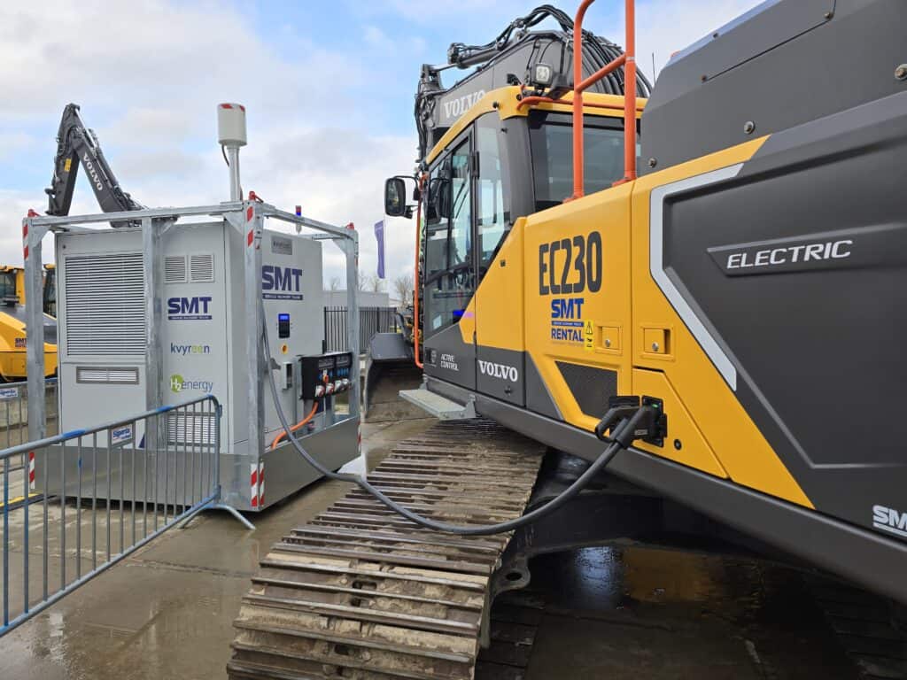 Une pelle électrique Volvo EC230 est connectée à une station de charge sur un chantier de construction, avec des câbles visibles et des barrières de sécurité.