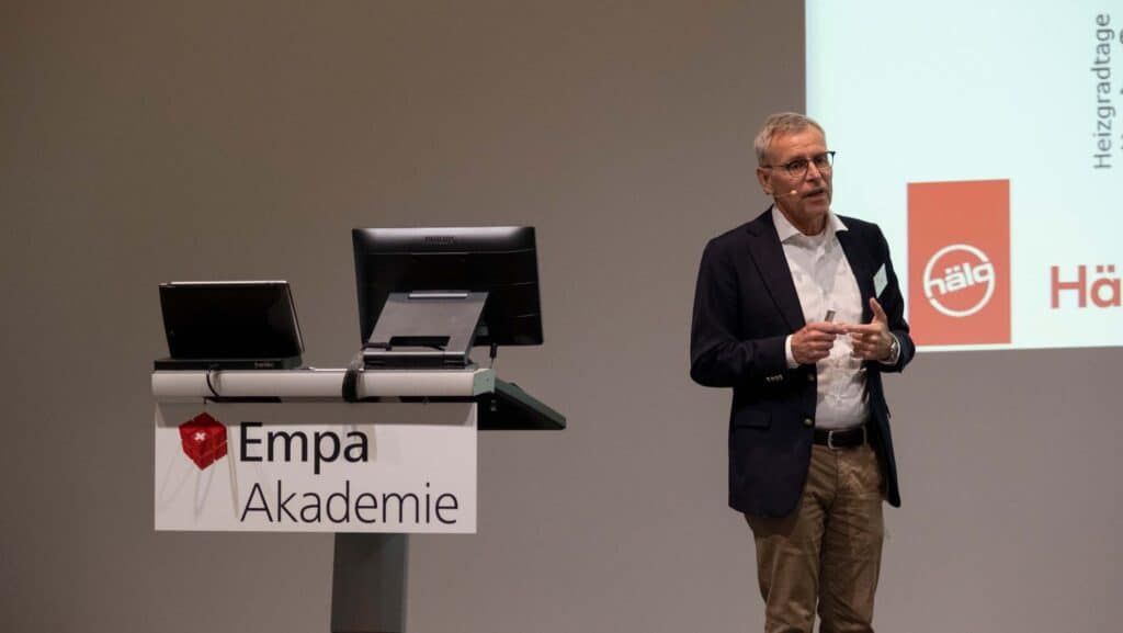 A man in a suit stands next to a podium labeled "Empa Akademie," giving a presentation with a projection screen behind him.