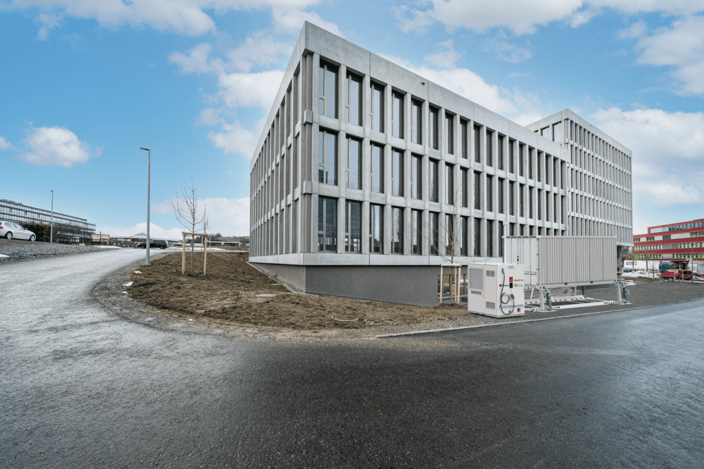 A modern, rectangular office building with tall vertical windows sits at a corner; a large industrial unit is installed outside near the road.
