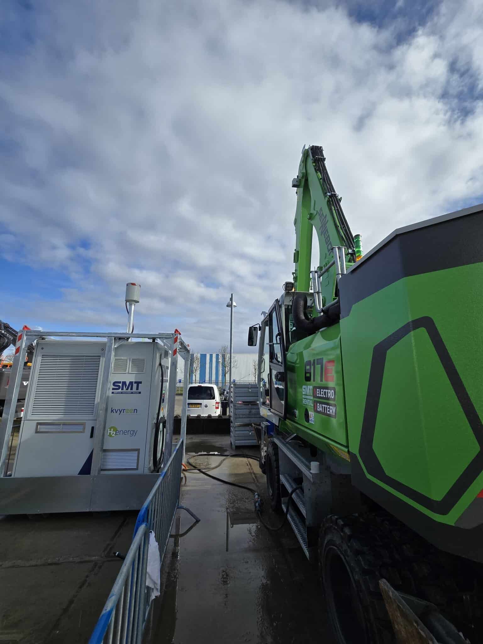 A green electric construction vehicle is parked beside a portable battery charging station on a cloudy day.