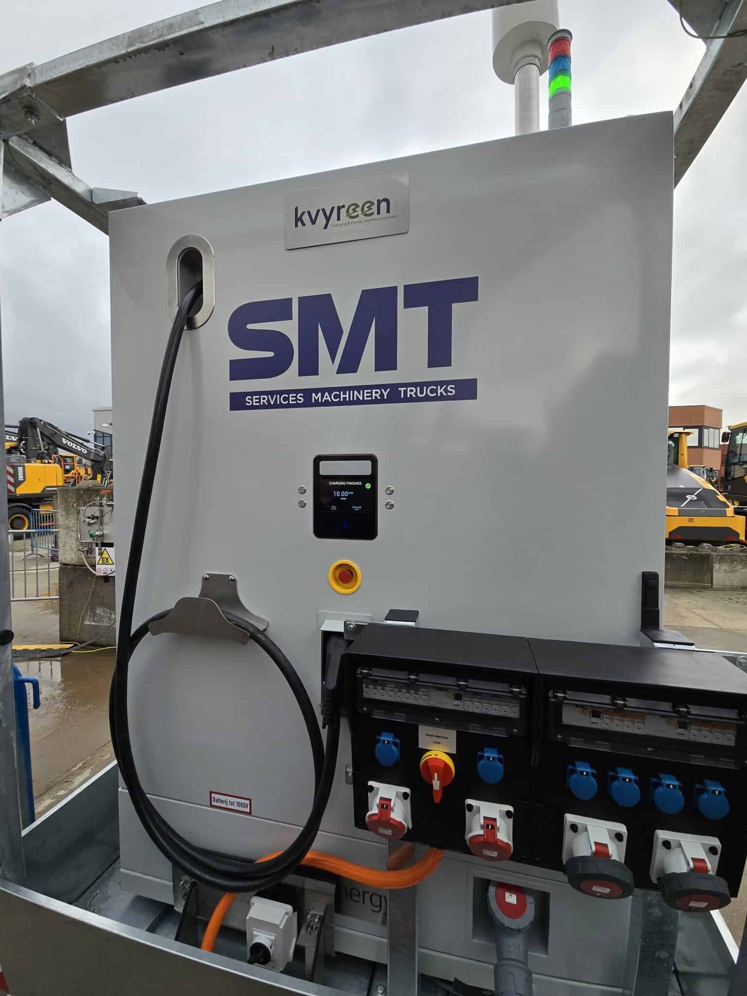 A charging or power distribution unit with the label "SMT Services Machinery Trucks," various sockets, cables, and control panels is shown outdoors.